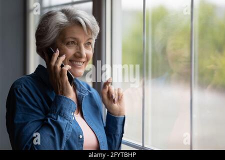 Femme plus âgée debout près de la fenêtre parlant sur un smartphone Banque D'Images