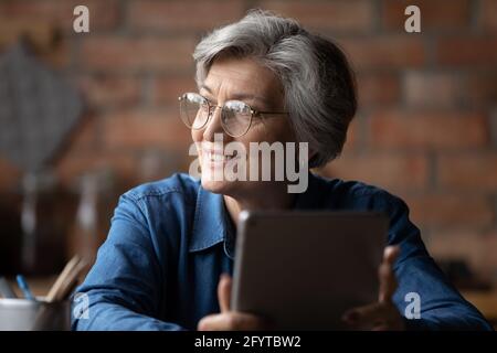 Jolie femme grise à cheveux assis à l'intérieur tenir la tablette en regardant loin Banque D'Images