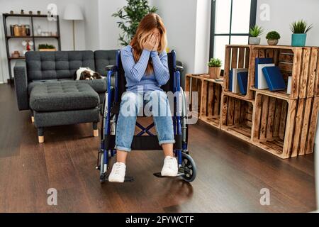 Jeune belle femme assise en fauteuil roulant à la maison avec une expression triste couvrant le visage avec les mains en pleurant. Concept de dépression. Banque D'Images