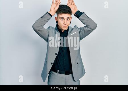 Jeune garçon caucasien avec des oreilles dilatation portant une veste d'affaires faisant des oreilles de lapin gestuelle avec les mains paumes ayant l'air cynique et sceptique. lapin de pâques Banque D'Images