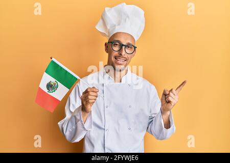 Homme chauve avec barbe portant un tablier de cuisinier professionnel tenant le mexique drapeau souriant heureux pointant avec la main et le doigt vers le côté Banque D'Images