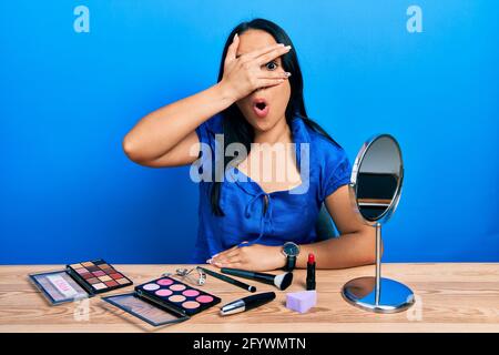 Belle femme hispanique avec le perçage du nez se préparer à l'aide de maquillage peeking dans le choc couvrant le visage et les yeux avec la main, regardant à travers les doigts afr Banque D'Images