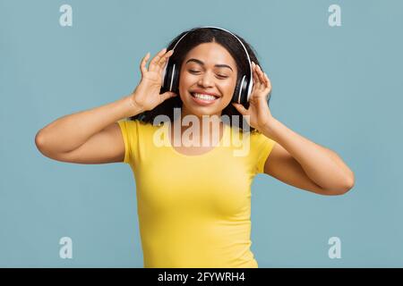 Une femme noire pleine de joie dans un casque sans fil écoutant de la musique avec des yeux fermés, fond bleu studio Banque D'Images