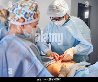 Chirurgien plastique et assistant effectuant la chirurgie plastique dans la salle d'opération, portant des masques médicaux et des uniformes chirurgicaux tout en enlevant l'excès de graisse de l'abdomen du patient. Concept d'abdominoplastie. Banque D'Images