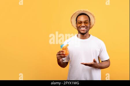 Happy Black Guy dans une tenue d'été offrant un verre de cocktail tropical sur fond jaune, bannière avec espace de copie Banque D'Images