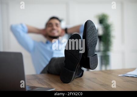 Un jeune employé arabe se détendant après un travail réussi, mettant des jambes sur table au bureau, attention sélective Banque D'Images