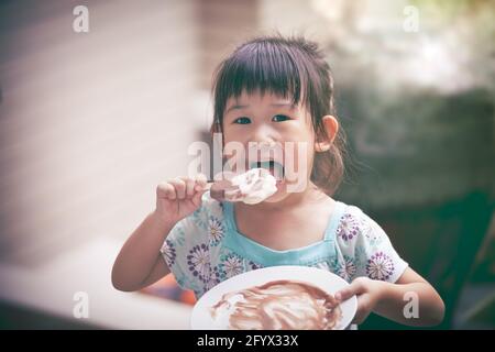 Jolie fille asiatique mangeant de la glace en été sur fond flou. Enfant heureux regardant l'appareil photo. À l'extérieur. Style vintage. Banque D'Images