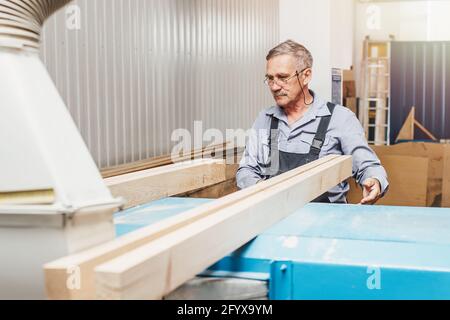 Portrait d'un menuisier âgé ou d'un charpentier en combinaison travaillant avec des planches en bois dans un atelier de menuisier. Sciage de bois. Banque D'Images