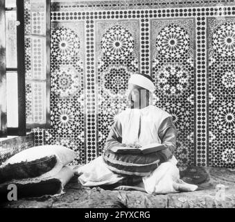 Un homme assis sur un tapis près d'une fenêtre ouverte lit le Coran, Fès, Maroc, 1923. (Photo de Burton Holmes) Banque D'Images