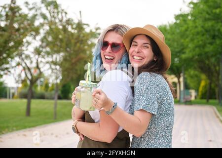 Deux amies joyeuses qui boivent des boissons rafraîchissantes, rient, se embrasent et regardent vers l'appareil photo dans un parc lors d'une journée ensoleillée d'été. Banque D'Images