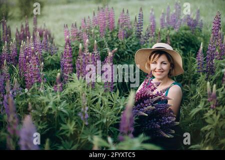 Une belle fille romantique sourit joyeusement dans une robe bleue et le chapeau est assis dans un champ de fleurs avec un énorme bouquet de fleurs lupin pourpres. Sélection douce Banque D'Images