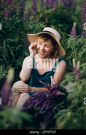 Une belle fille romantique sourit joyeusement dans une robe bleue et le chapeau est assis dans un champ de fleurs lupin pourpres. Mise au point sélective douce. Banque D'Images