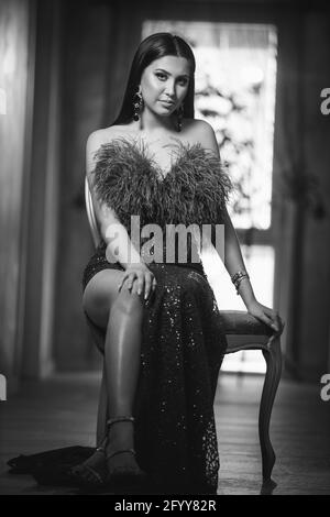 Photo verticale en niveaux de gris d'une jeune femme brune posant assis sur une chaise dans une robe de soirée Banque D'Images