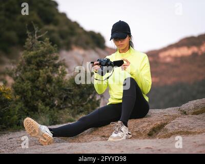 Jeune femme hispanique en activité et Cap regardant la vidéo sur un caméscope assis sur une falaise rocheuse pendant le trekking dans la nature vallée Banque D'Images