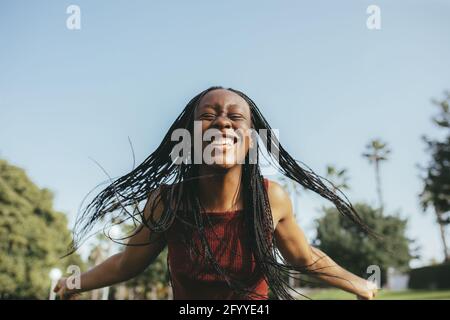 Joyeuse femme afro-américaine aux yeux fermés et aux cheveux volants s'amuser dans la rue avec des arbres verts jour d'été Banque D'Images