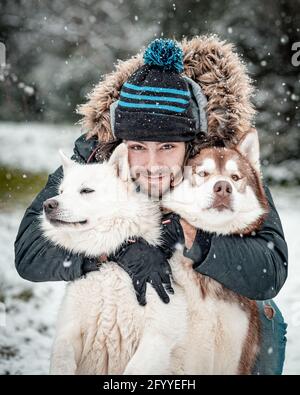 Jeune homme positif dans des vêtements chauds embrassant l'adorable Husky et Berger suisse blanc tout en passant du temps ensemble dans les bois d'hiver en chute de neige Banque D'Images