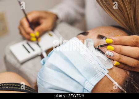 Rognez un cosmétologue méconnaissable à l'aide d'une pince à épiler en appliquant de faux cils pour l'extension à l'œil du client dans le salon Banque D'Images