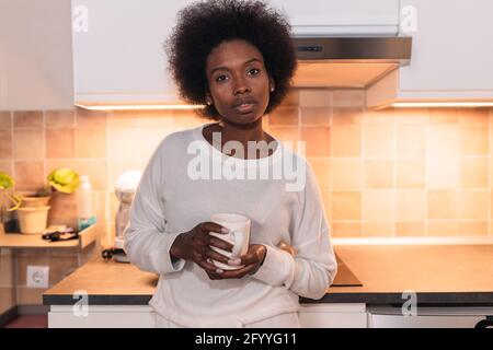 Jeune femme ethnique tranquille avec des cheveux afro sombres dans une ambiance décontractée préparer une tasse de café chaud et regarder l'appareil photo en vous tenant près de la table dans la salle de cuisine Banque D'Images