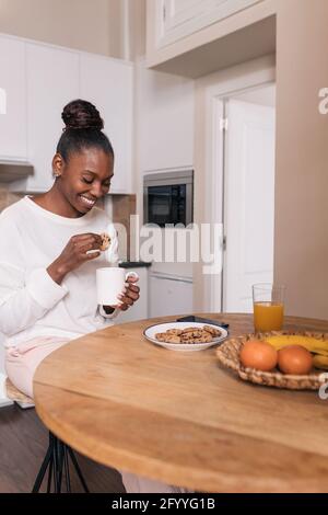 Positive jeune femme noire avec des cheveux foncés dans des vêtements décontractés en trempant des biscuits dans une tasse de café tout en prenant le petit déjeuner dans la cuisine Banque D'Images