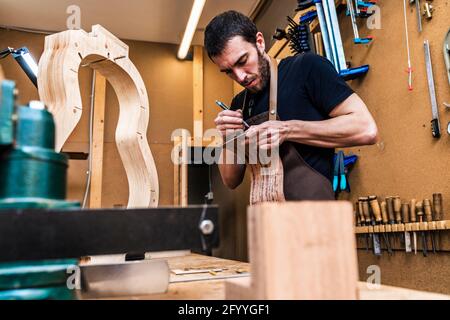 L'artisan mâle barbu écrit sur une pièce en bois tout en faisant de la musique instrument dans la salle de travail avec divers outils manuels Banque D'Images