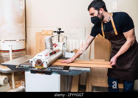 Vue latérale d'un jeune homme artisan portant un masque de protection mise à niveau de pièces en bois avec machine de surfaçage avec tube en atelier pendant le covid 19 pand Banque D'Images