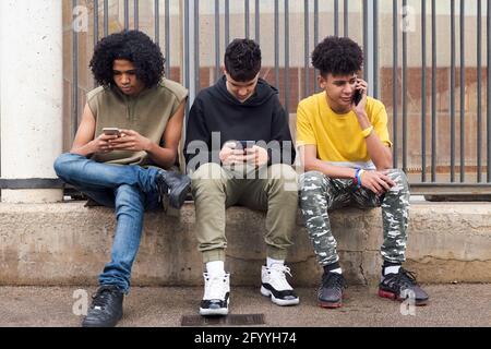 Des amis adolescents multiraciaux et sérieux surfant sur Internet sur des téléphones portables assis contre la clôture métallique et en regardant l'écran Banque D'Images