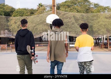 Vue arrière des adolescents multiraciaux de sexe masculin avec basketball et planches à roulettes debout ensemble contre le filet de sport dans le parc Banque D'Images
