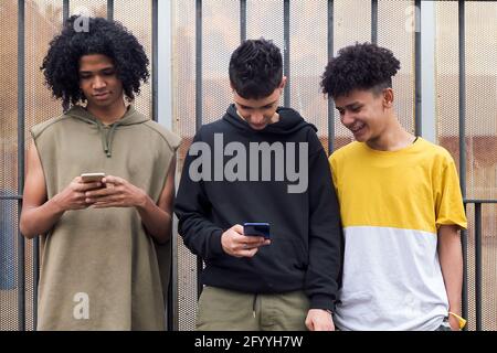 Des amis adolescents multiraciaux surfant sur Internet sur des téléphones mobiles se tenant contre clôture en métal et regard à l'écran Banque D'Images
