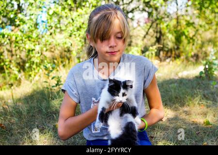 Flou blonde petite fille souriante jouant avec chat, noir et blanc petit chaton. Nature flou vert d'été arrière-plan. Fille tenant et de stroking p Banque D'Images