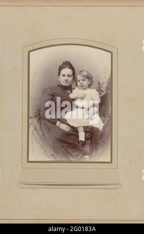 Studio portrait de la mère et de la fille. Studio portrait de la mère et de la fille. La mère porte une robe noire à col fermé avec une broche. La fille porte une robe blanche, ensemble ils s'assoient sur une chaise. Banque D'Images