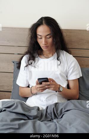Sérieux jeune homme vietnamien avec de longs cheveux noirs assis sous couette grise dans le lit et message texte sur le smartphone Banque D'Images