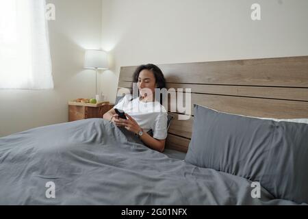 Contenu beau jeune homme vietnamien avec des cheveux longs noirs couchés dans le lit avec lampe commutée et utiliser le téléphone tout en gaspillant heure le matin Banque D'Images