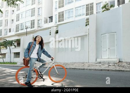 Sérieux élégant jeune vietnamien gars en denim porté vélo avec des roues orange contre un bâtiment moderne Banque D'Images