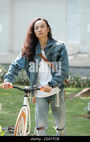 Portrait d'un jeune asiatique sérieux et beau avec sac épaule tenant des poignées de vélo tout en marchant dans le parc d'été Banque D'Images