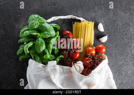 Sac réutilisable avec articles d'épicerie. Sac fourre-tout, gaspillage minimal. Basilic frais, tomates cerises, ail dans un sac de tissu sur fond de table sombre. Vue de dessus, copier la flèche Banque D'Images