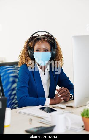Portrait d'une jeune femme d'affaires gaie dans un casque et un masque médical salon au bureau devant l'ordinateur et vue à l'appareil photo Banque D'Images