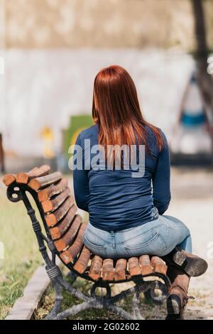 Une femme aux cheveux rouges assise sur un banc avec son dos à la caméra un jour ensoleillé. Tir américain. Banque D'Images