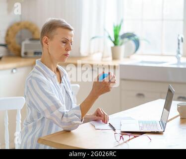 Femme mûre focalisée s'exerçant avec une bague de préhension ronde en caoutchouc pour les paumes pendant le travail ou les études sur ordinateur portable, prévention du syndrome de tunnel et le réchauffement de la main. Concept de technologies et de personnes âgées Banque D'Images