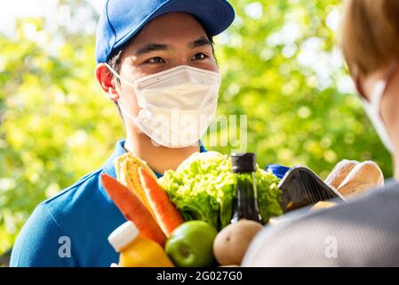 Un livreur asiatique hygiénique portant un masque facial tout en livrant des produits d'épicerie au client à la maison, livraison de nourriture en temps de pandémie concept Banque D'Images
