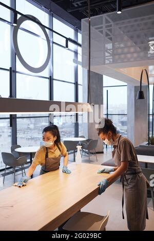 Portrait vertical de deux travailleuses portant des masques tout en nettoyant les tables dans l'intérieur moderne du restaurant, concept de sécurité covid, espace de copie Banque D'Images