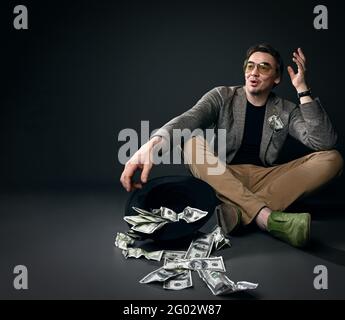 Homme d'âge moyen en veste, jeans et chaussures assis sur le sol par chapeau à l'envers plein de dollars d'argent liquide, raconter des histoires Banque D'Images