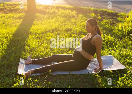 Paisible jeune femme enceinte positive en costume de gymnastique fait du yoga et médite assis sur un tapis d'herbe verte le jour chaud ensoleillé d'été. Concept de Banque D'Images