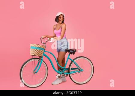 Charmante jeune femme noire en été porter debout avec un vélo classique et regardant l'espace vide, fond rose Banque D'Images