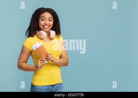 Concept de déplacement. Joyeuse femme afro-américaine avec écouteurs tenant des billets sur fond bleu studio, espace libre Banque D'Images
