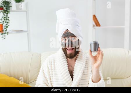 Homme barbu s'amusant avec un masque cosmétique sur son visage en argile noire. Hommes soins de la peau, humour et spa à la maison concept Banque D'Images