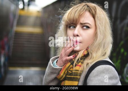 Jeune femme blonde sérieuse fume une cigarette et regarde de côté dans le passage souterrain Banque D'Images