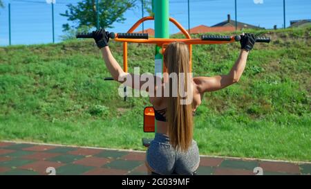 Jeune femme méconnaissable pratiquant une large poignée de traction à l'aide d'un simulateur sur le terrain de sport en plein air. Vue arrière de la fillette en forme avec corps musculaire parfait et entraînement de queue de cheval dans le matin ensoleillé. Banque D'Images