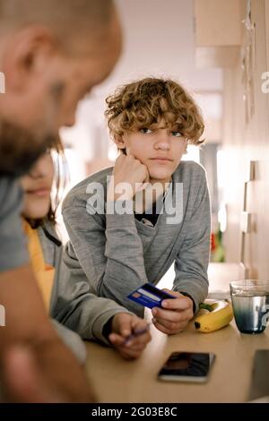 Garçon avec une carte de crédit regardant le père en se tenant à l'île de cuisine Banque D'Images