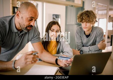 Frère et soeur regardant le père expliquant la finance avec carte de crédit à l'île de cuisine Banque D'Images