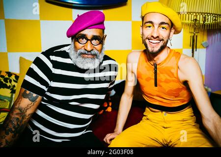 Jeune homme souriant avec un ami masculin senior portant des bérets assis dans la salle de séjour Banque D'Images